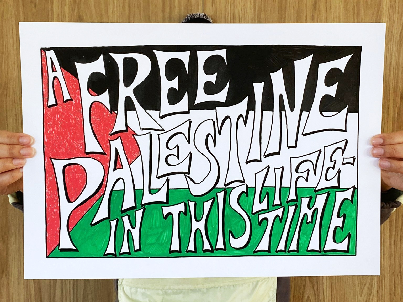  a person holding up a landscape A3 handdrawn poster. The poster depicts the Palestinian flag. Written across it in spiky lettering are the words: A free Palestine in this lifetime.