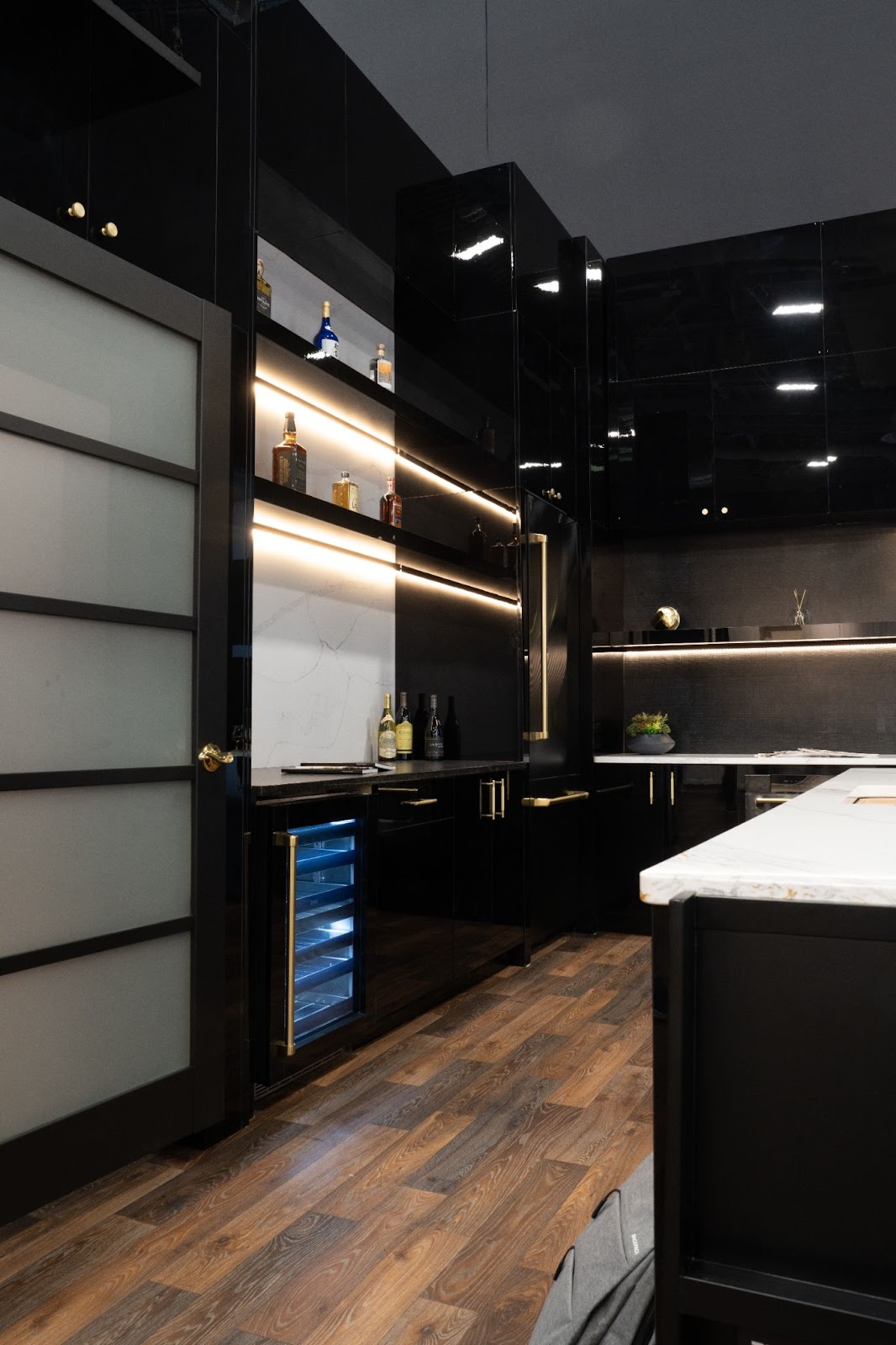 Modern kitchen design featuring high-gloss black cabinetry, gold accents, and under-cabinet lighting. A Touchstone built-in wine cooler is visible, alongside a variety of bottles on open shelving, set against a herringbone-patterned wooden floor.