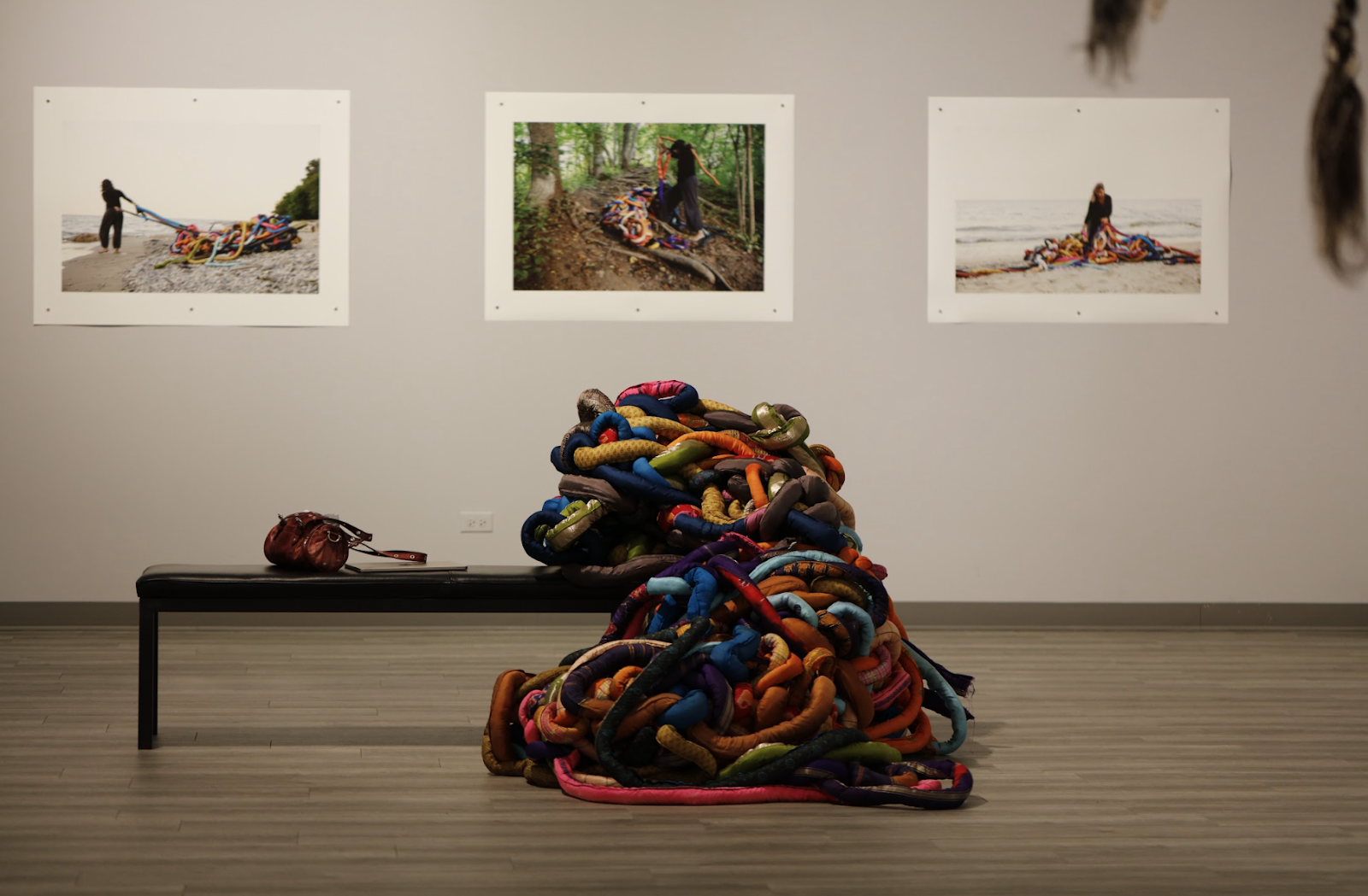 Image: Installation view, Entangled (2015). On the floor in the foreground, is a bench with a small leather bag on it. Further to the right of the bag are many multi-colored, thick tubes of fabric, tangled and knotted thickly around each other. The Weight of Our Past (2021), photographs, hung on the wall behind it. Image courtesy of the South Asia Institute.