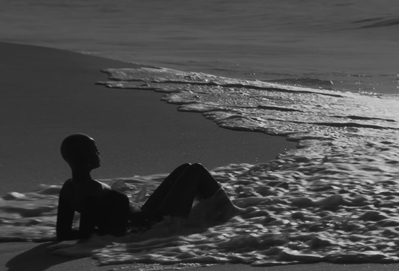 A desaturated screenshot of Kelela from her music video for "Enough for Love". Featuring a shot of her laying on a beach, as the tide comes in and washes over her.