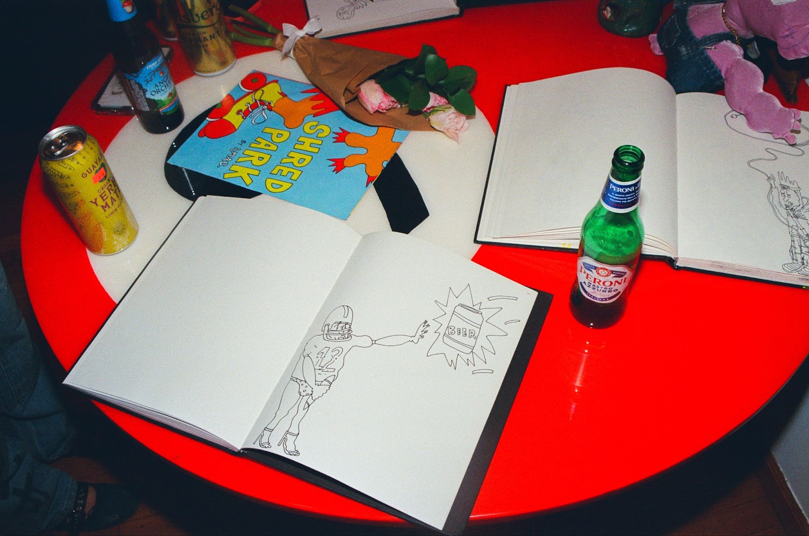 Alt text: A circular red table with two white notebooks, a zine, a bouquet of flowers, and a couple of bottles.