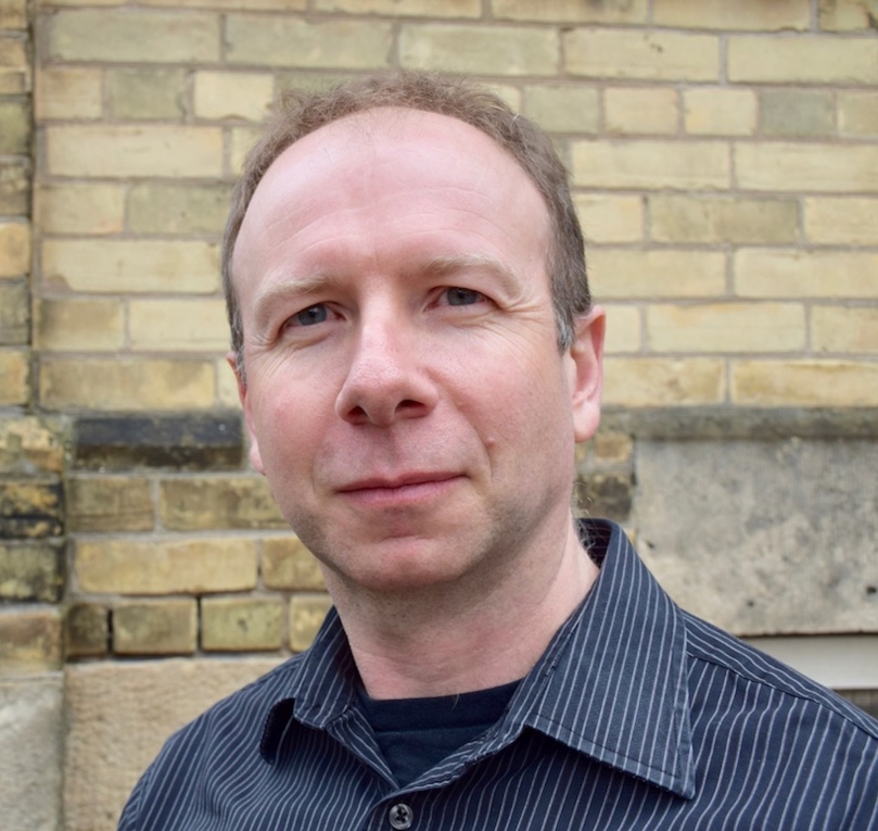A headshot of Dr. Kieran O'Doherty, PhD who is wearing a black pinstripe shirt and standing outside in front of a yellow brick wall.