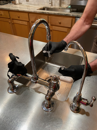 A scientist wearing black gloves is collecting a sample of tap water from the kitchen sink using small plastic vials. 