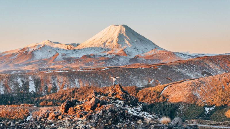 A person with arms outstretched stands in front of the snow-covered peaks of Tongariro National Park at sunrise.