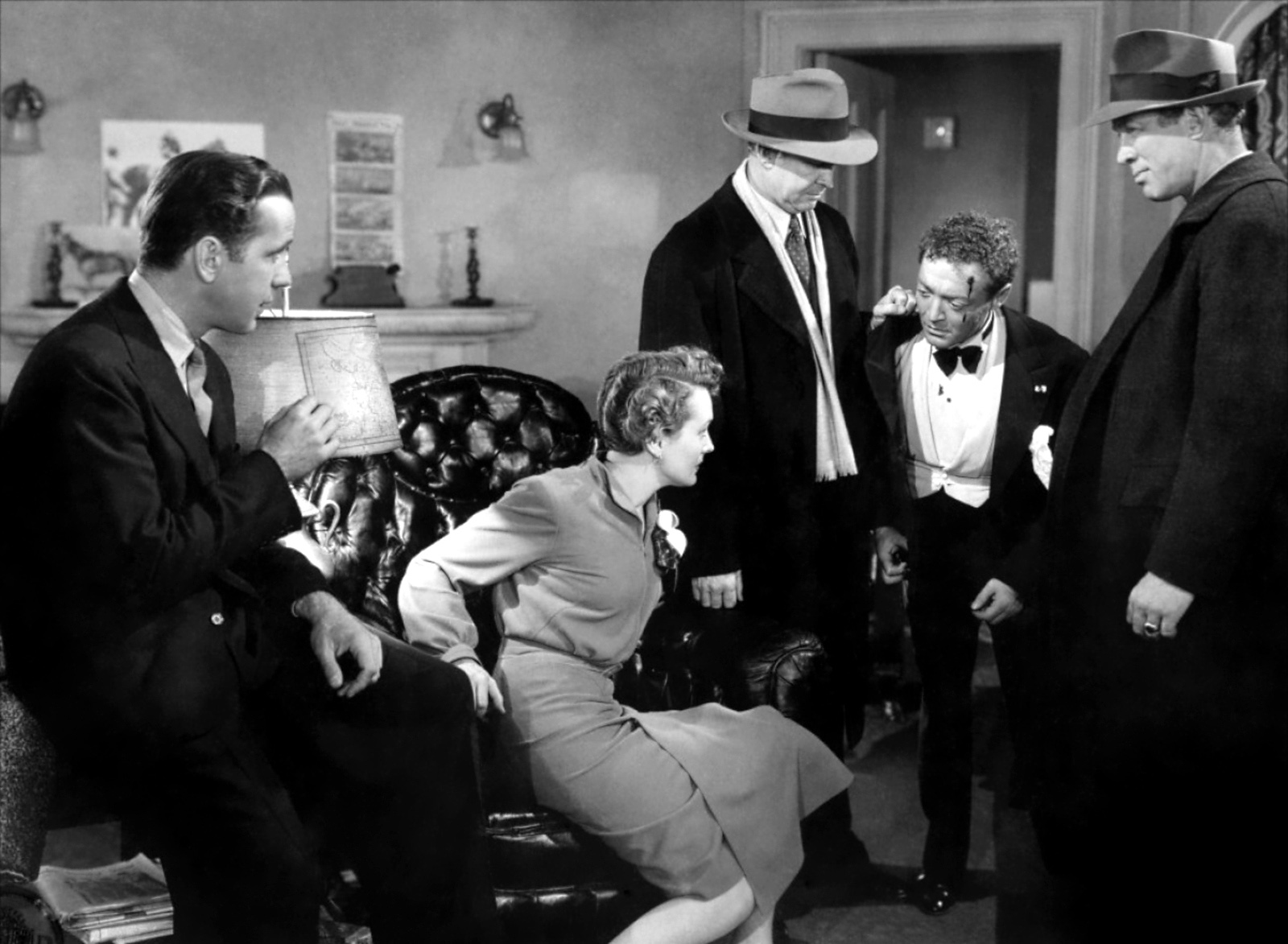 Four men in suits, two in felt fedoras, sit and stand around the room. One man on the right has a bleeding head wound. A woman sits in a leather chair in the middle of the scene looking at the wounded man. Black and White photograph. 