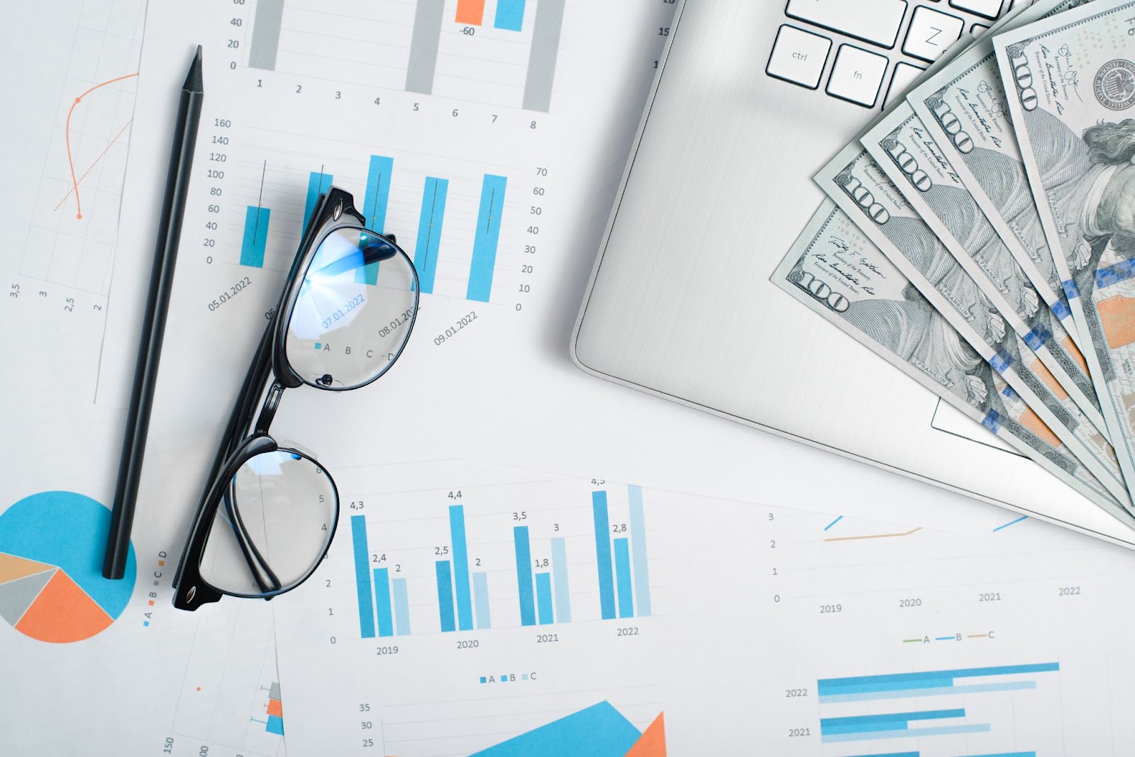 A top view of a table displaying business documents, a laptop, dollar bills, eyeglasses, and a pencil illustrating a business financial review. 