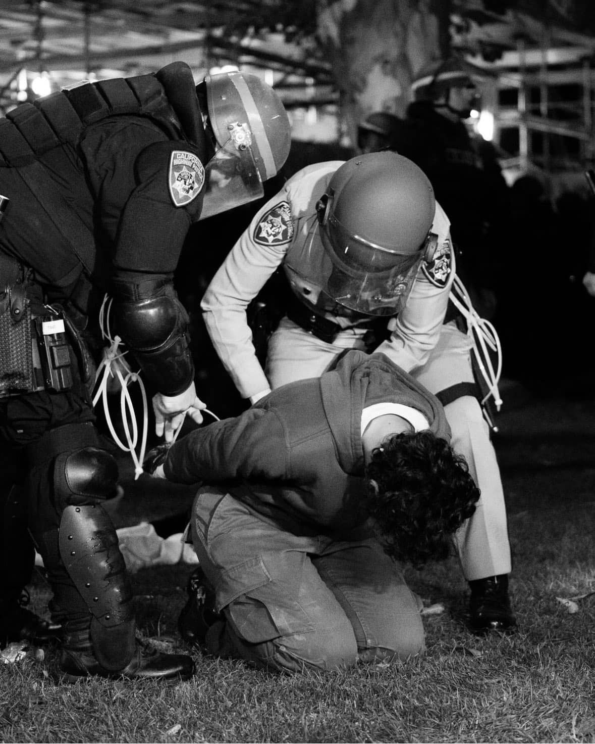 A student kneels down, face lowered, as two riot police handcuff their hands behind their back.