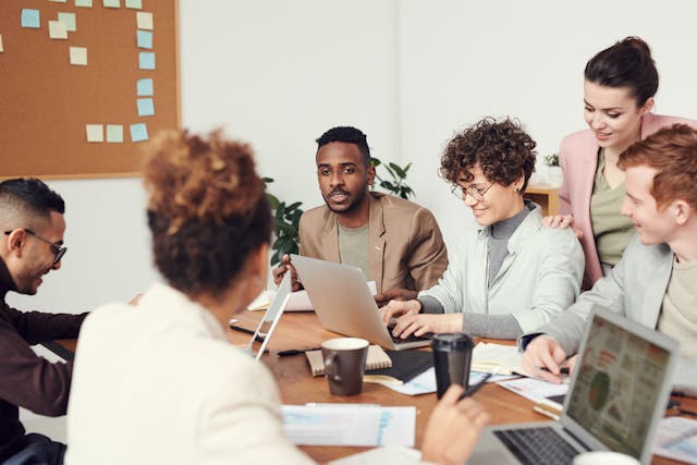 A group of professionals sitting around a table with laptops, papers, and coffee cups during a meeting in an office setting.