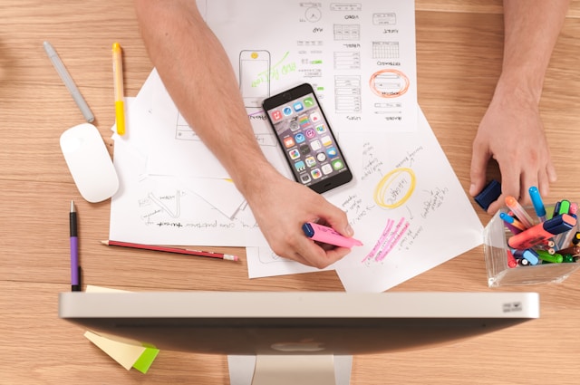 A wooden table with papers and a monitor used by a UI UX designer to sketch a wireframe