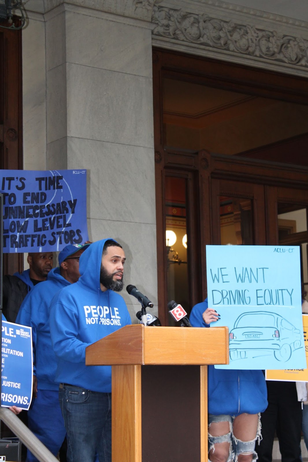 An ACLU of CT Smart Justice leader speaks at a rally at the capitol building in Hartford, Connecticut. He stands in front of signs that say "We want driving equity" and "It's time to end unnecessary low level traffic stops" and other, similar signage.