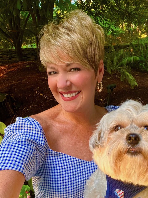 With her garden as the backdrop, Cindy wears a blue and white gingham patterned blouse and holds her puppy.
