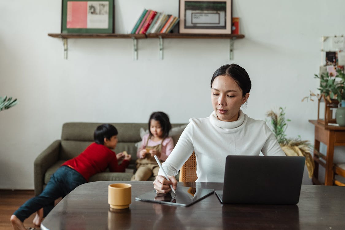 Free Focused young Asian mother working remotely while children playing on sofa Stock Photo