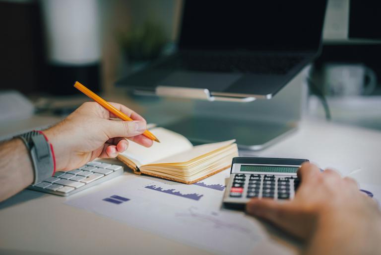 A man with a pencil, notebook, and calculator in front of his laptop while planning their company's budget to book an athlete speaker.