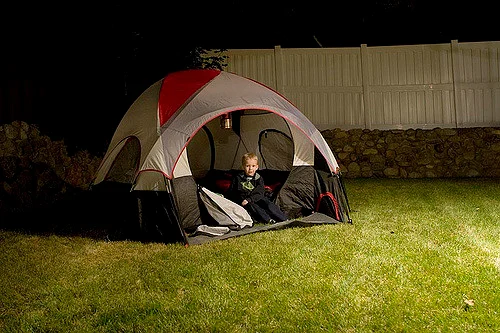 A small boy sits in a tent in a fenced backyard.