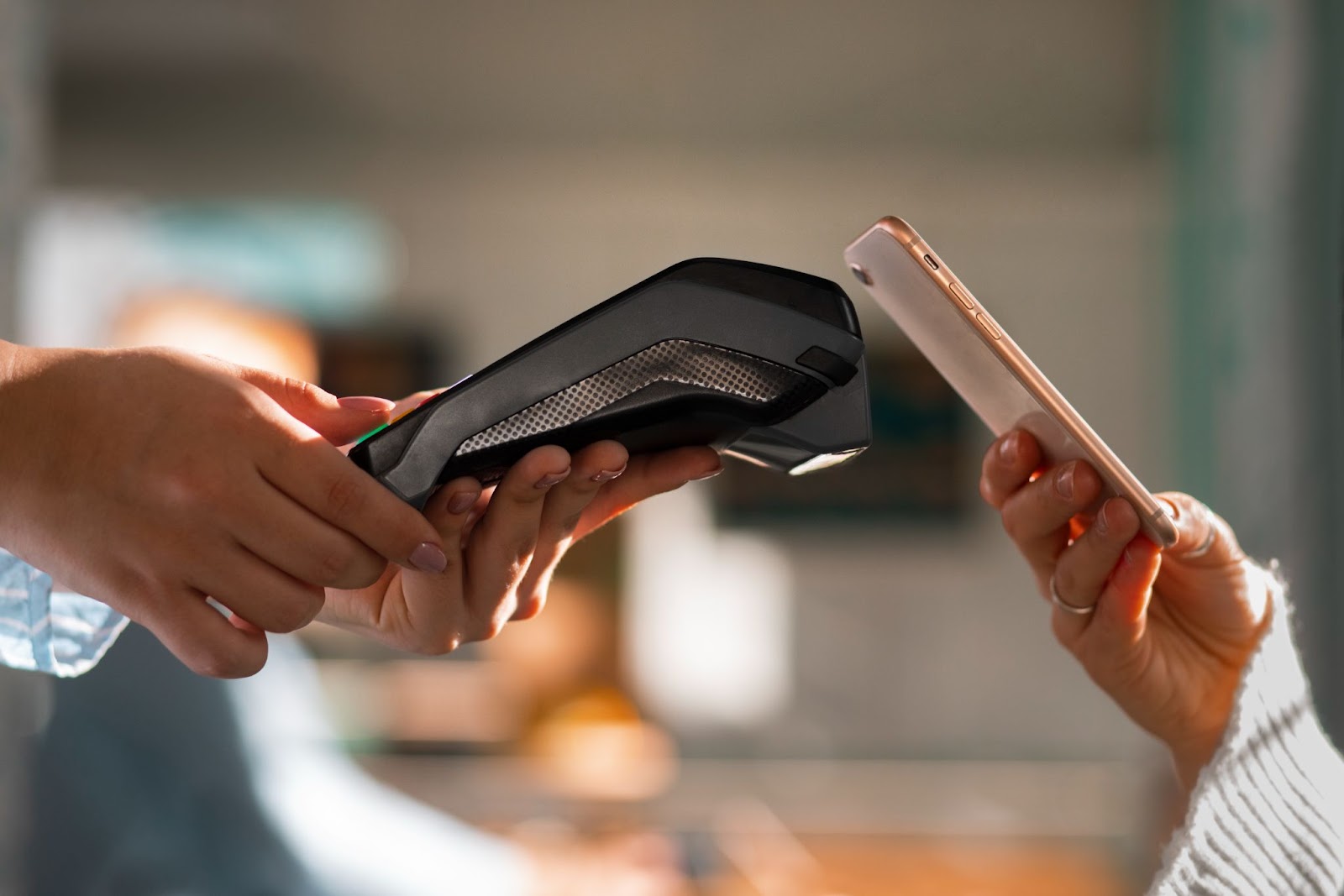 Imagem de uma maquininha de cartão e um celular sendo encostado próximo, fazendo um pagamento por aproximação. As mãos que seguram os objetos parecem femininas.
