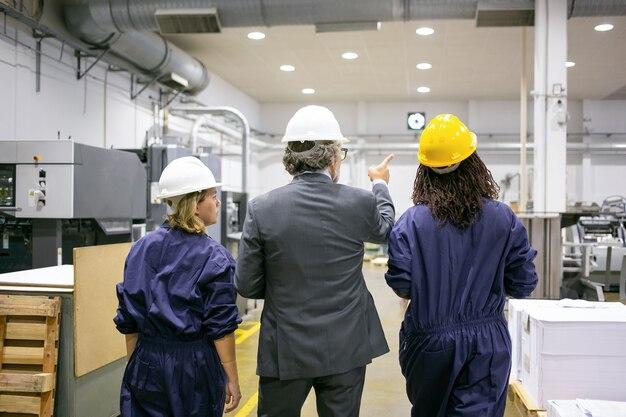 Free photo male engineer and female factory employees in hardhats walking on plant floor and talking, man pointing at equipment and instructing women