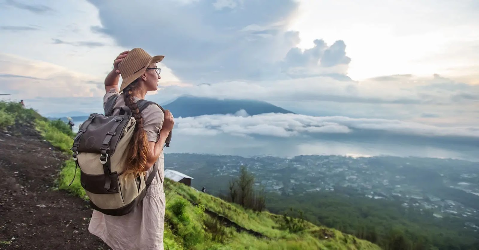 A woman, seen from behind, with her hair in a braid and wearing a beige sunhat and a grey backpack, stands on a mountain path overlooking a sea of clouds with mountains in the distance, portraying a sense of adventure and tranquility.