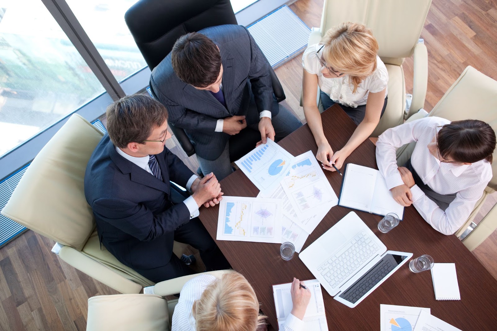 A top view of a business meeting with two male and three female professionals seated around a table, discussing a report displayed on documents and a laptop.