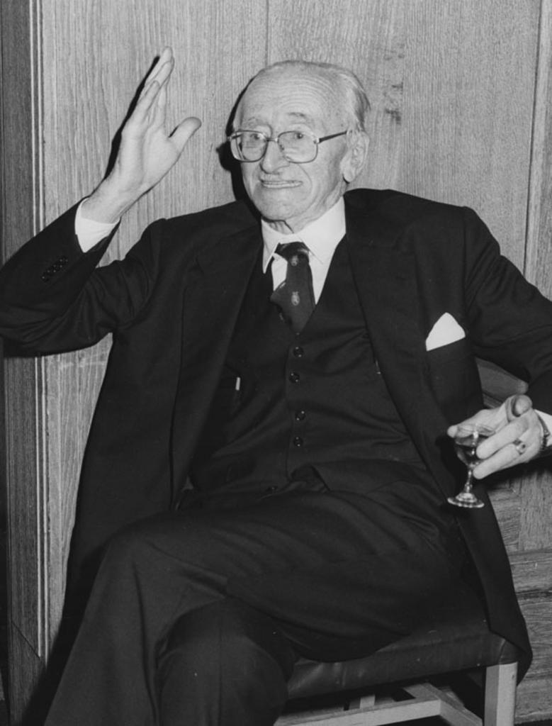 Friedrich Hayek is wearing a dark suit and tie. He is sitting in a chair. One hand holds a small sherry glass and the other is gesturing emphatically. 