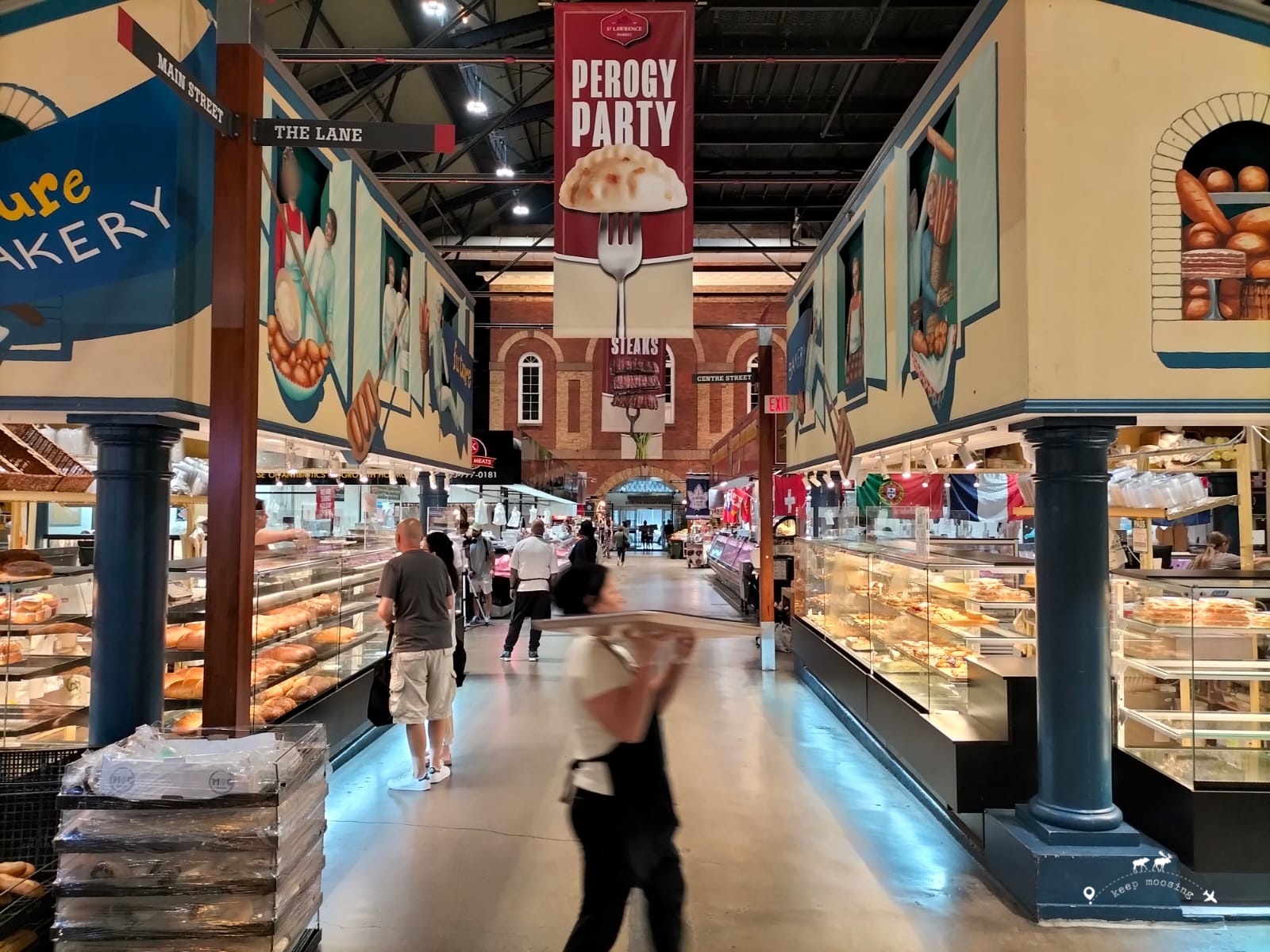 The interior of the St. Lawrence Market with a baker in the foreground carrying a tray of bread. The benches at the sides of the image.