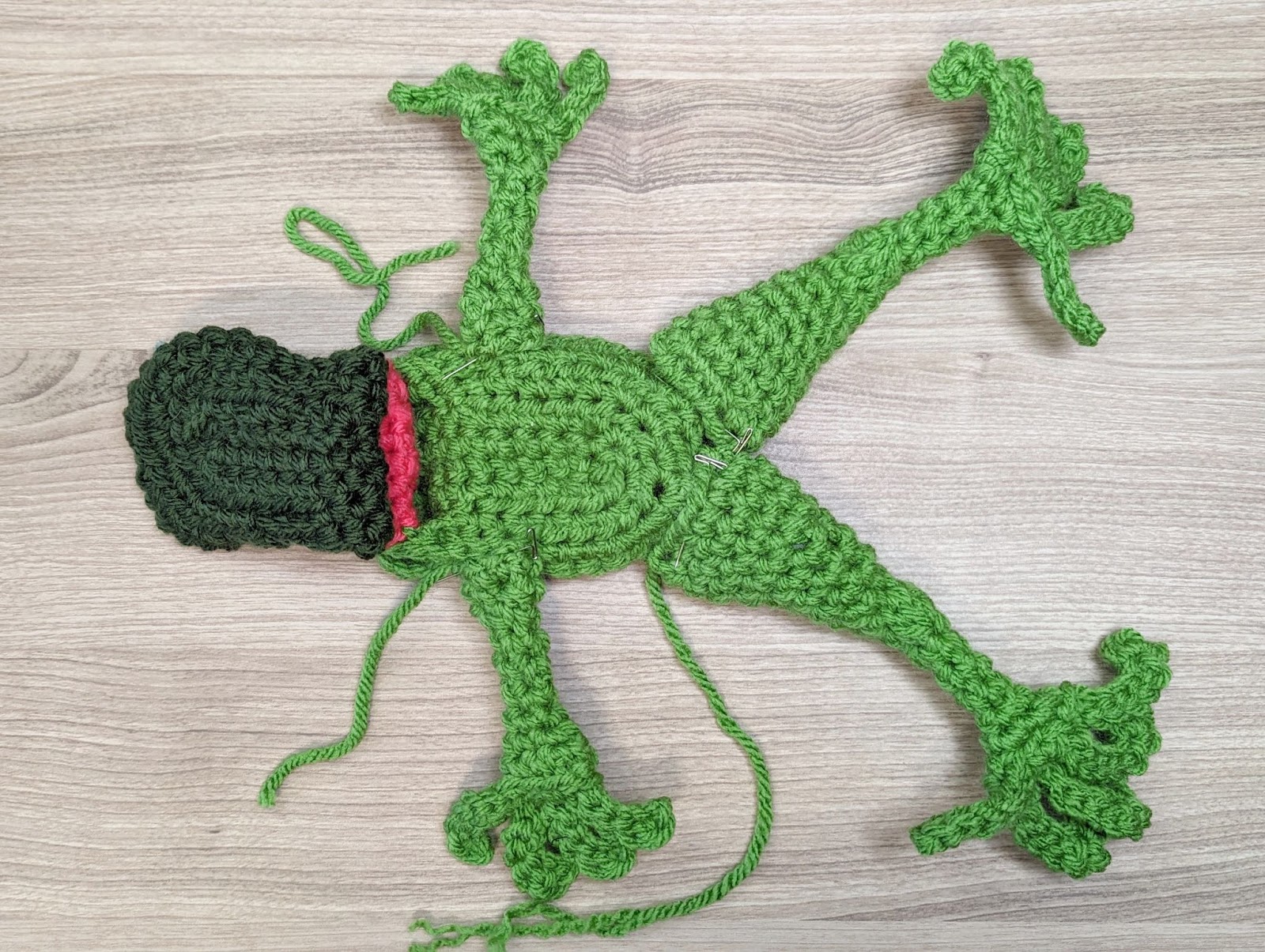 A handmade, green crocheted frog is laid out flat on a wooden surface. The frog's limbs are spread out and it has a red mouth with some loose green yarn strands around its body. The texture of the crochet is clearly visible.