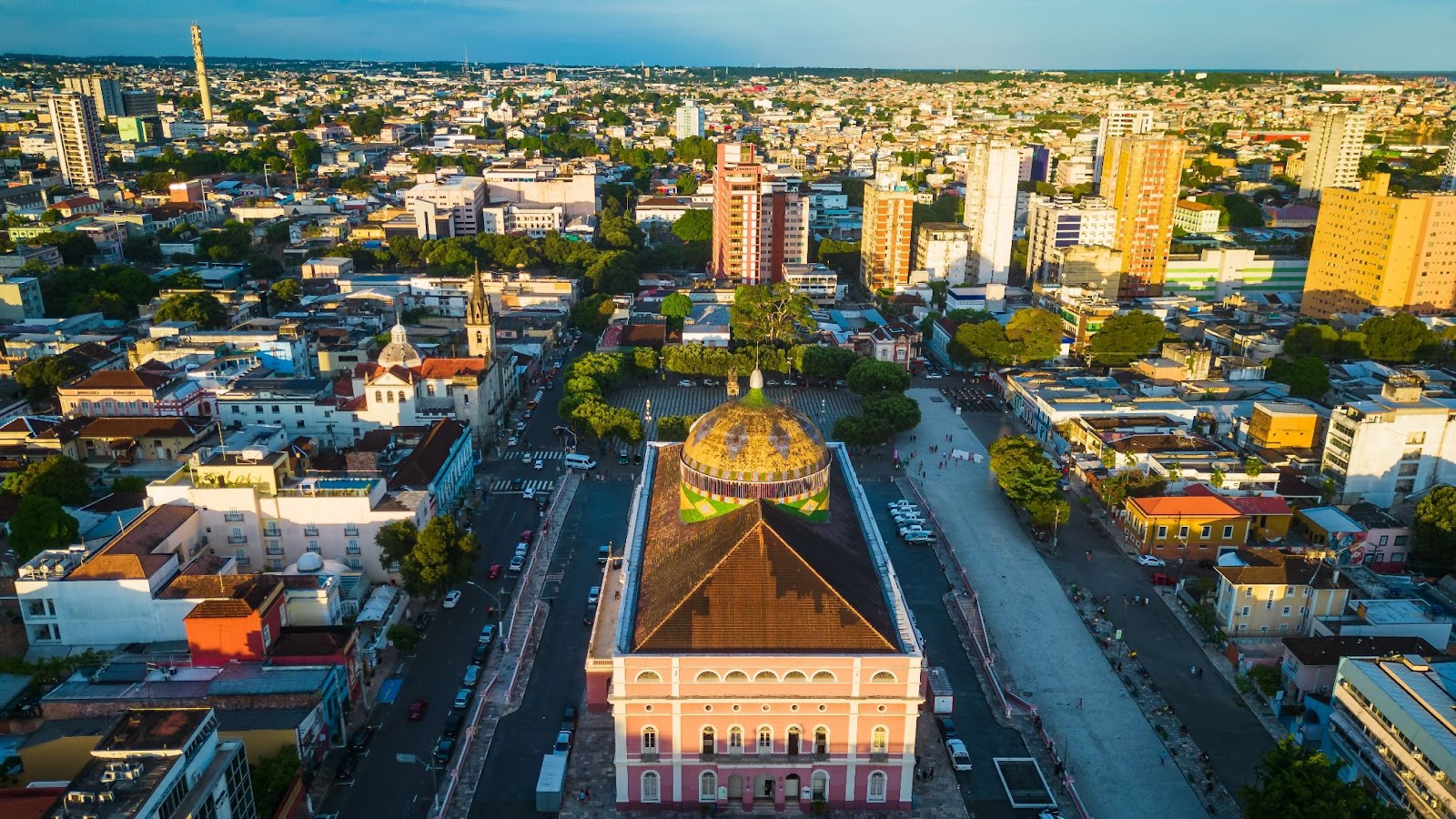 Vista aérea panorâmica da cidade de Manaus ao entardecer. Diversos prédios pelas ruas arborizadas.