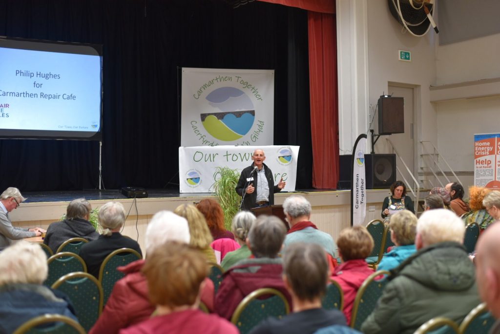 A man with a microphone stands in front of a room packed with people, introducing his project - Carmarthern Repair Cafe. 