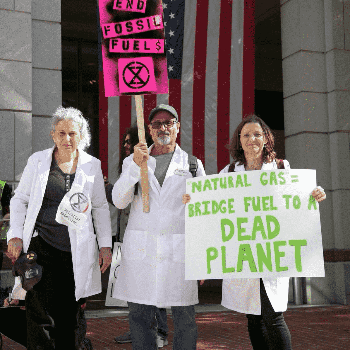 3 scientists stand before an American flag with anti-fossil fuel signs.