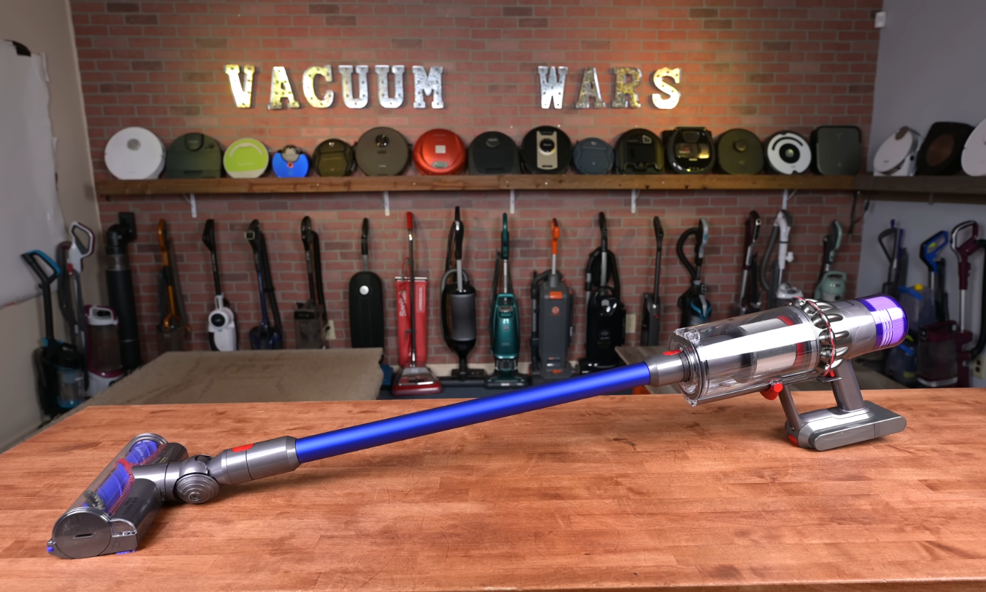 The Dyson V11 cordless vacuum displayed prominently on a wooden table in a well-organized workshop with various vacuum cleaners showcased in the background