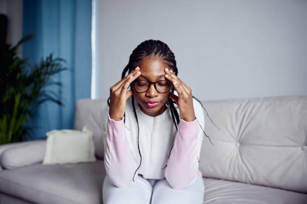 a young girl is sitting on the couch at home with her head in her hands. - black woman stress stock pictures, royalty-free photos & images