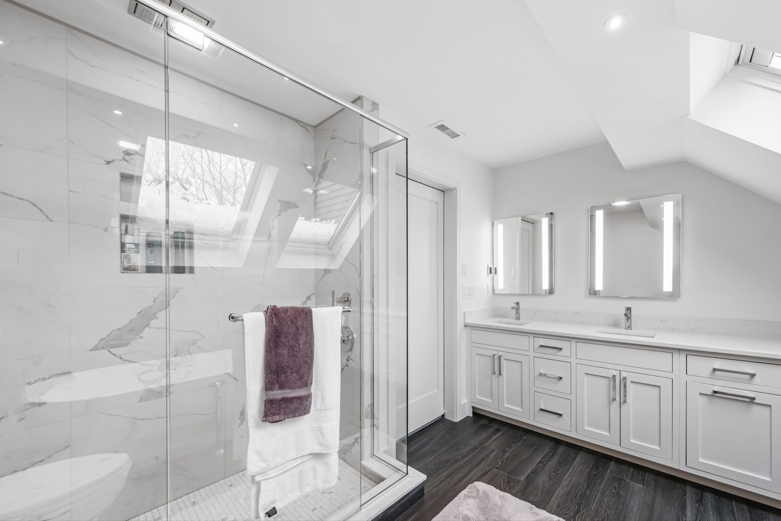 A modern white bathroom with glass partition, dual sinks, and mirrors with crisp white and brown towels hanging elegantly on the door handle.
