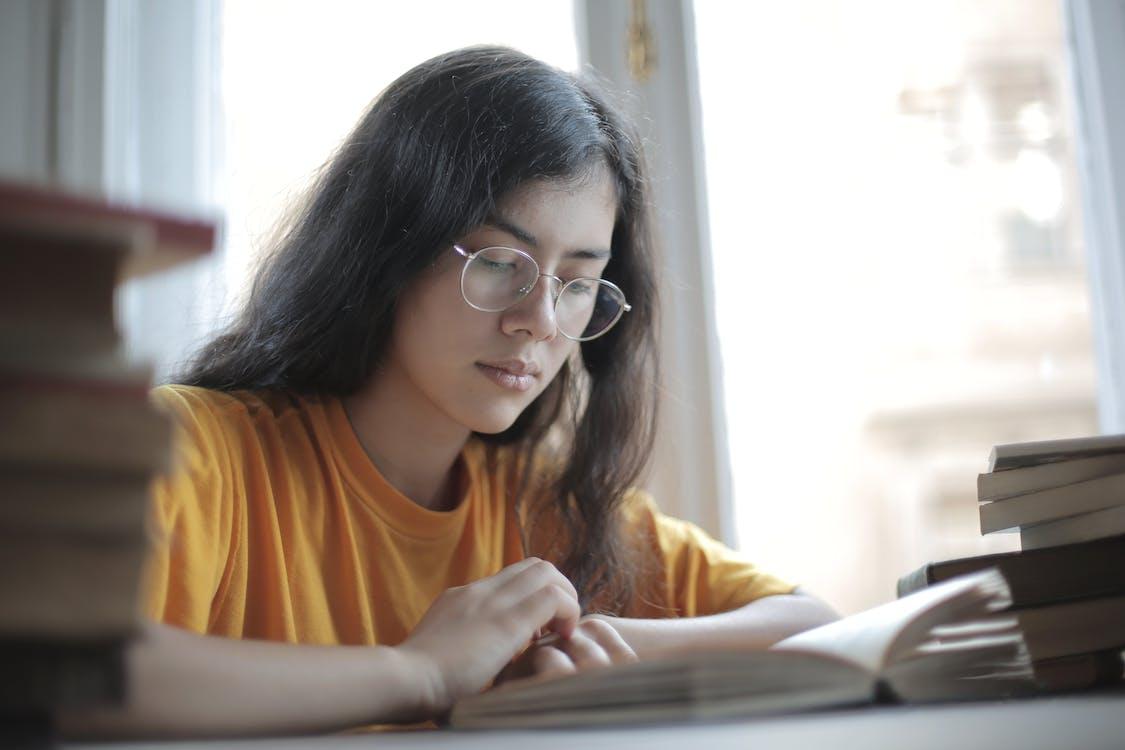 Free Low angle of diligent ethnic female student wearing casual t shirt and eyeglasses sitting at table with stacks of books and preparing for exam Stock Photo
