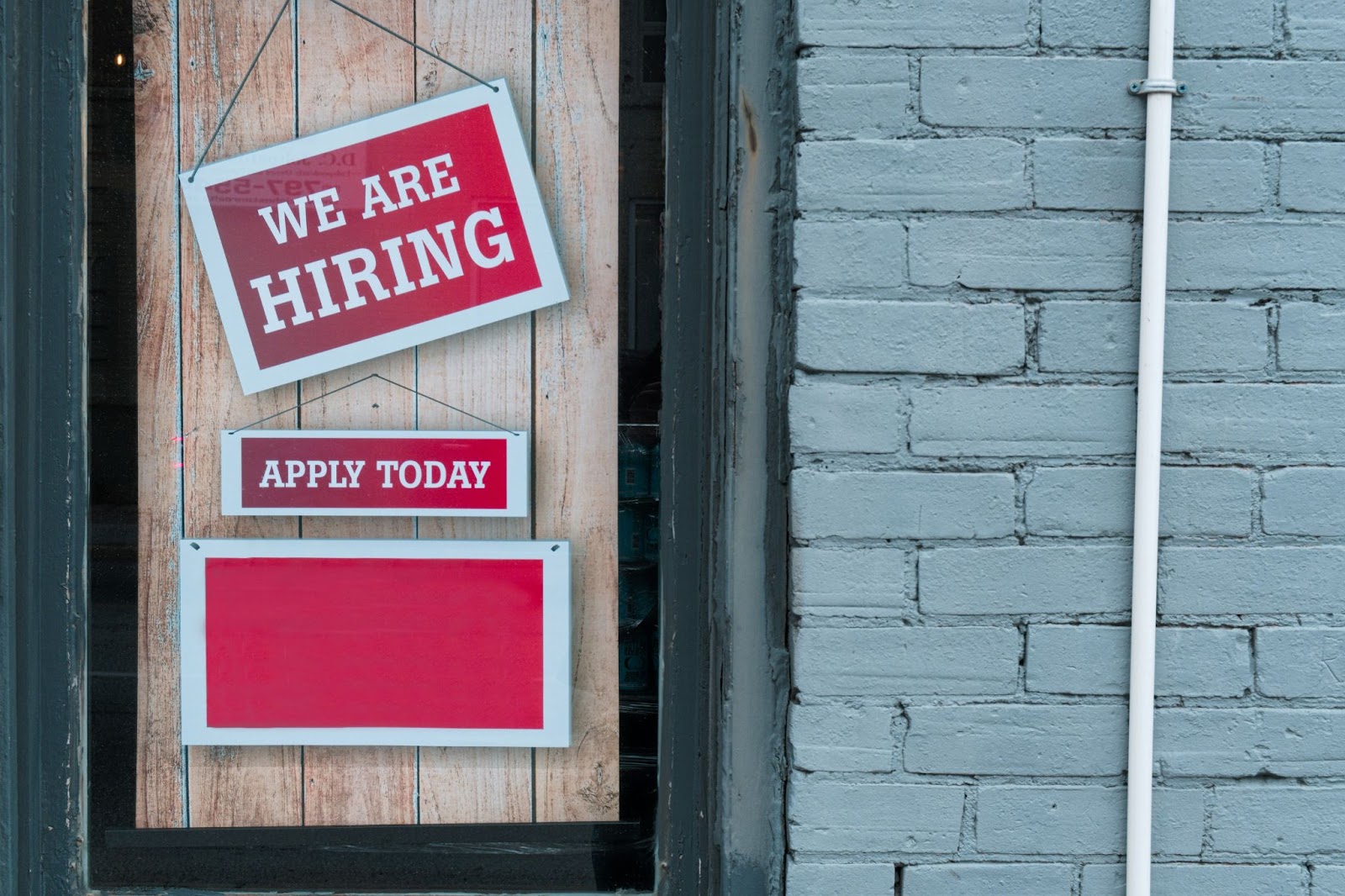 We are hiring & apply today signs hanging on a door next to a gray, painted, brick wall.