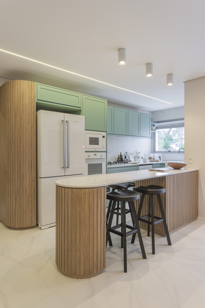 Cozinha planejada em madeira e móveis verde claro, com bancada branca, bancos pretos e geladeira branca.