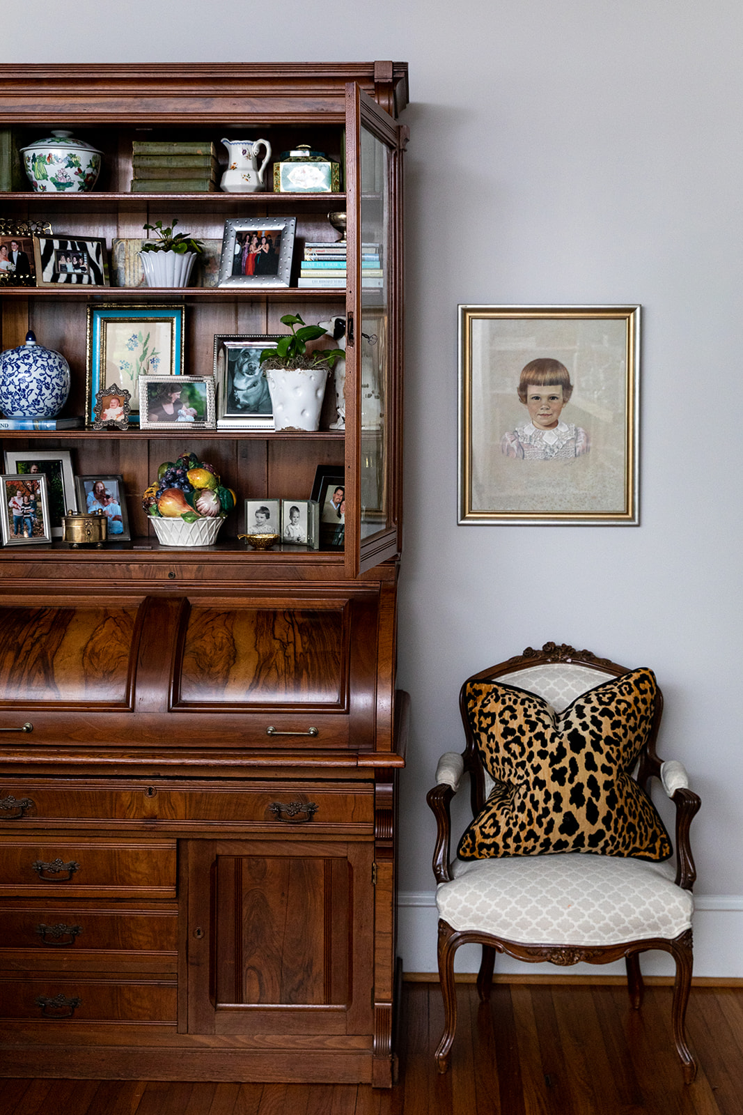 Brown wood hutch decorated with books, image frames, porcelain jars, urns and vases with a touch of greenery and fruit. Next to it staged a baby girl painting and an armchair with a leopard pattern throw pillow.