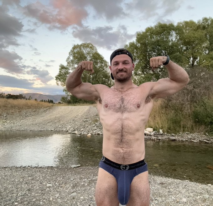 Max Small flexing his biceps standing outside near a creek wearing nothing but a pair of navy trunk underwear