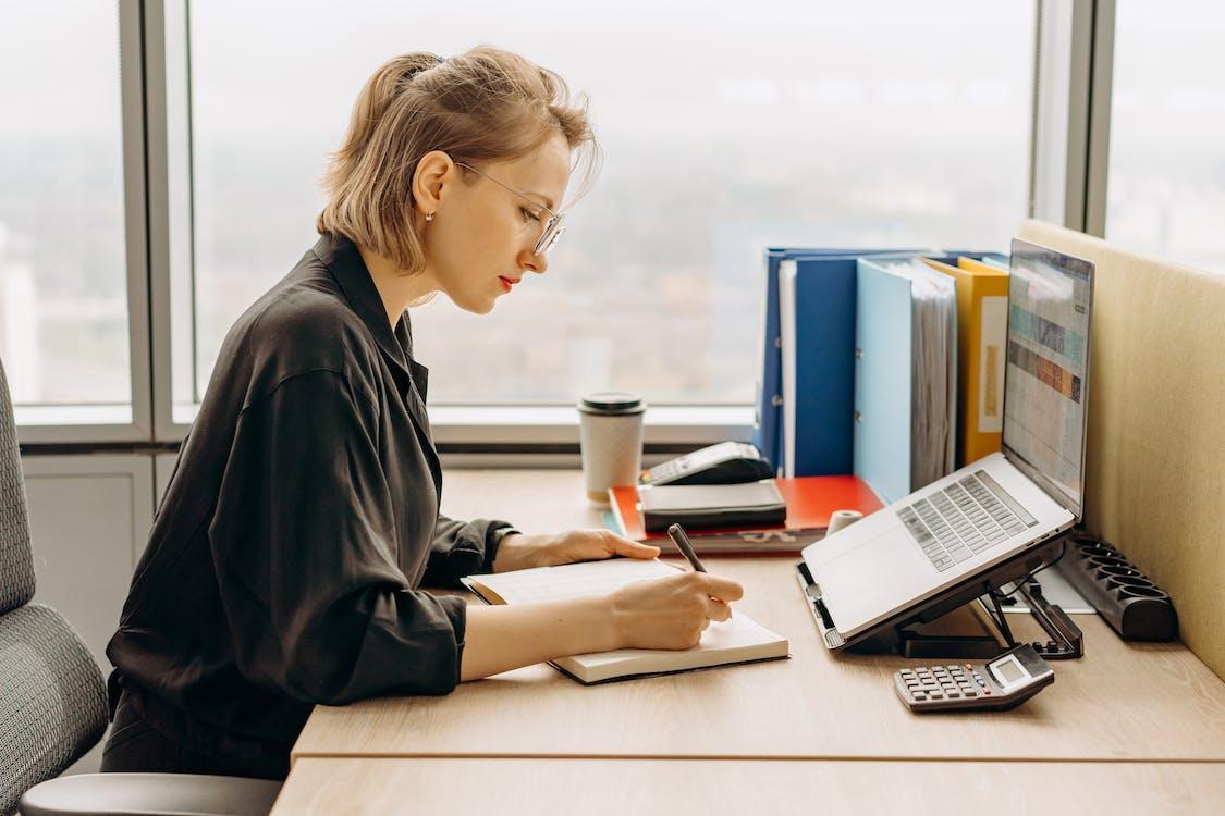 Free Woman in Black Long Sleeve Shirt Writing on a Notebook Near a Laptop Stock Photo