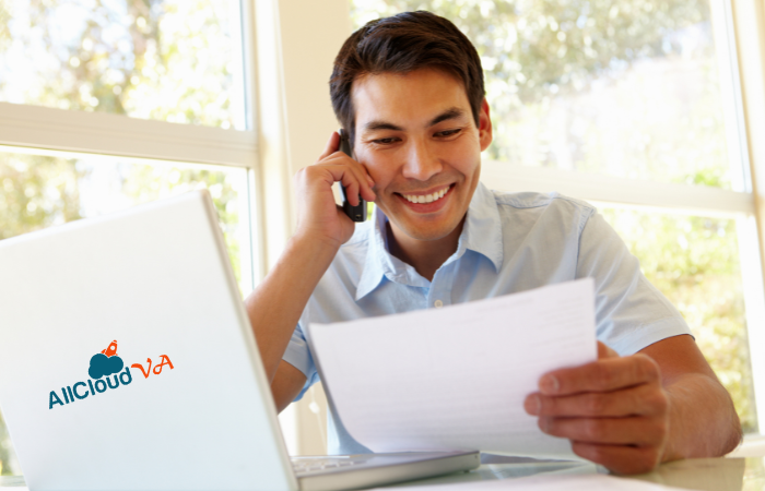 A cheerful man on the phone with documents and a laptop featuring the "AllCloud VA" logo, likely assisting a client.