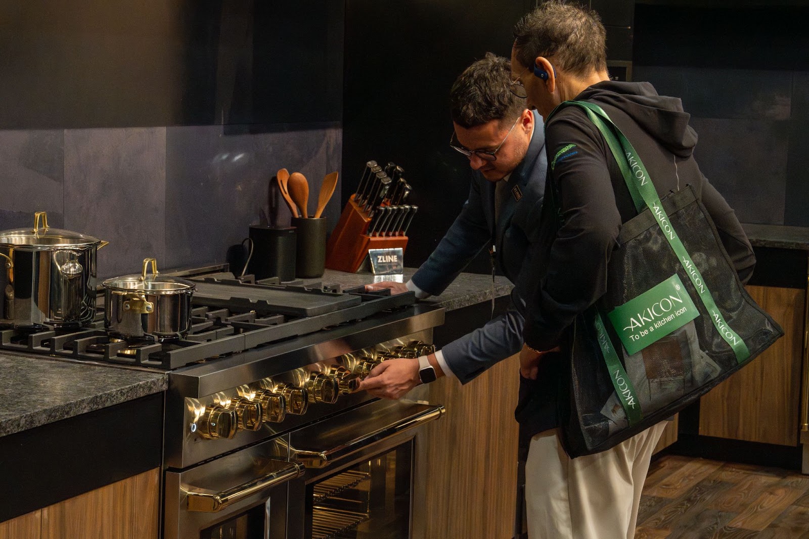 Two individuals inspecting a professional gas range (ZLINE's SGR48) with brass details at a trade show. The stove is accompanied by stainless steel cookware and a set of knives, against a backdrop of dark kitchen cabinetry.