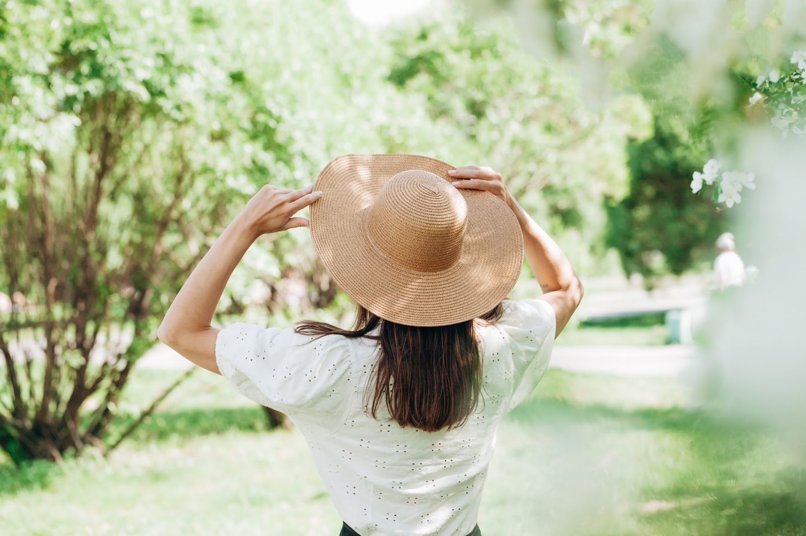 Mulher de costas com chapéu de palha e camiseta branca, observando as árvores e o gramado de um parque.