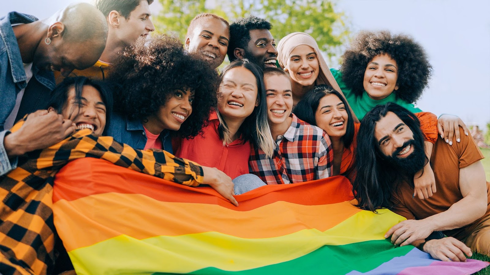 Grupo de pessoas de diferentes idades, etnias, gêneros e estilos sorrindo enquanto seguram uma bandeira do orgulho LGBT+.