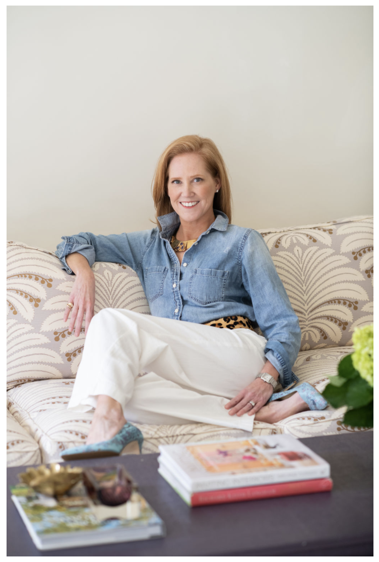 Lindsey is wearing a denim shirt and white pants as she sits on a patterned sofa in a styled living room vignette.