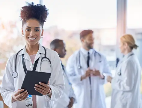 Woman Doctor With Stethoscope Around Neck and Tablet in Hand With Her Colleagues in The Background