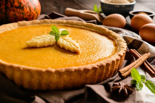 A whole pumpkin pie surrounded by cinnamon sticks, eggs, mint leaves, and star anise