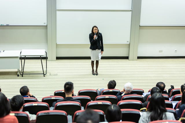 A woman standing at the front of a lecture hall speaking to an audience of seated people.