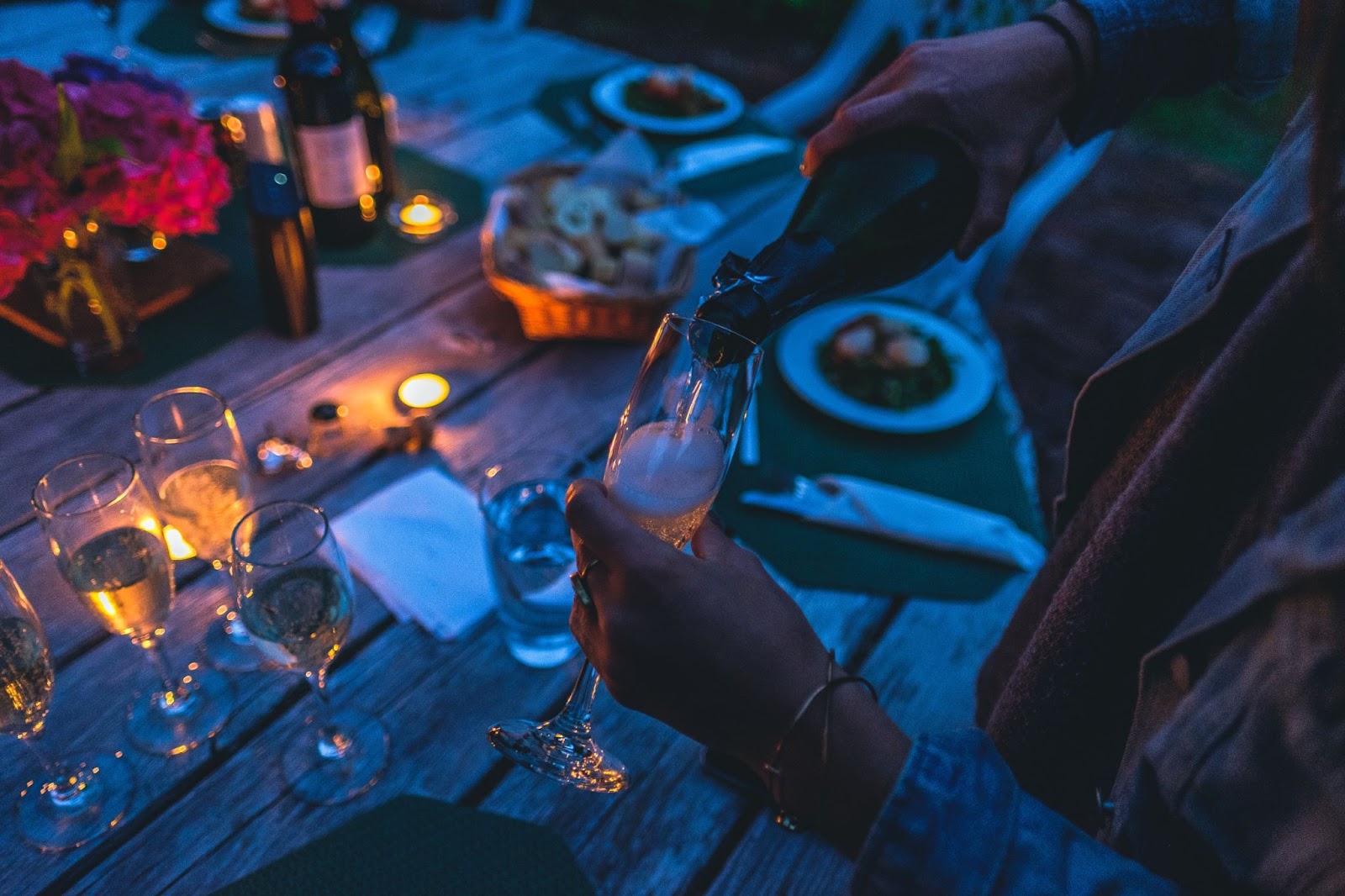 A person pours sparkling wine into a flute glass at a dimly lit outdoor dining table adorned with candles, creating a cozy and inviting atmosphere.