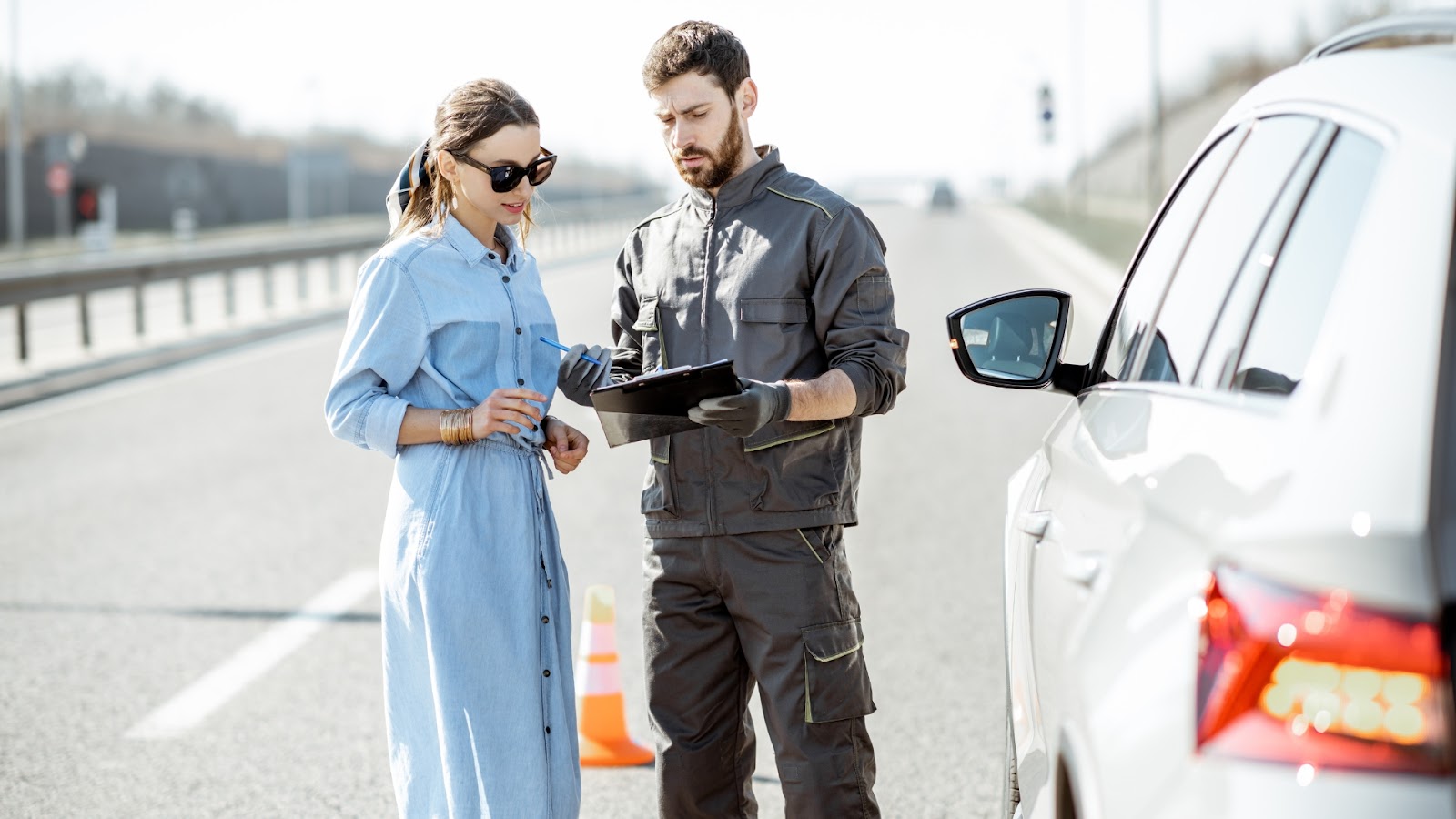 A roadside assistance professional provides car lockout service to a female motorist stranded on a highway.