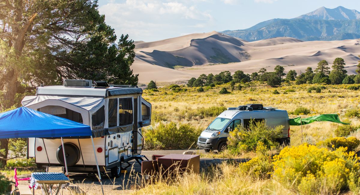 RV camping in scenic Colorado with a pop-up camper and a van parked beside lush greenery and wildflowers, with majestic Great Sand Dunes and mountain peaks in the background under a clear blue sky