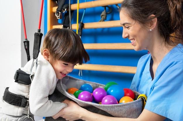 Foto niño lindo con parálisis cerebral haciendo terapia musculoesquelética en el hospital mientras se ríe y se divierte ...