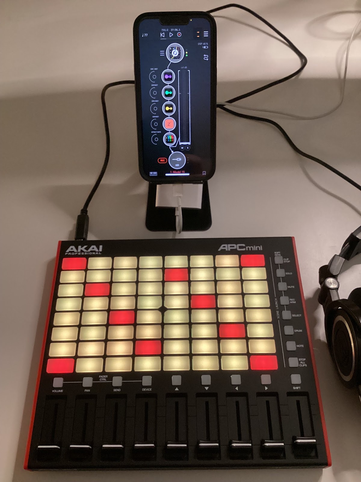 An iPhone running AUM with five effects plugins connected to an Akai APC Mini with its keys lit up in red and yellow/green, next to a pair of headphones.